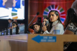 Woman speaking into a microphone, with SDG wheel on the wall, looking like a halo