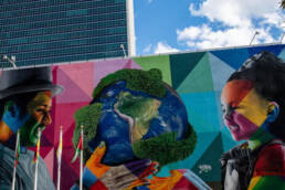 A photo of a UN wall that has art on it. A man holding a globe, passing it to a young girl. Lots of colours and geometric shapes